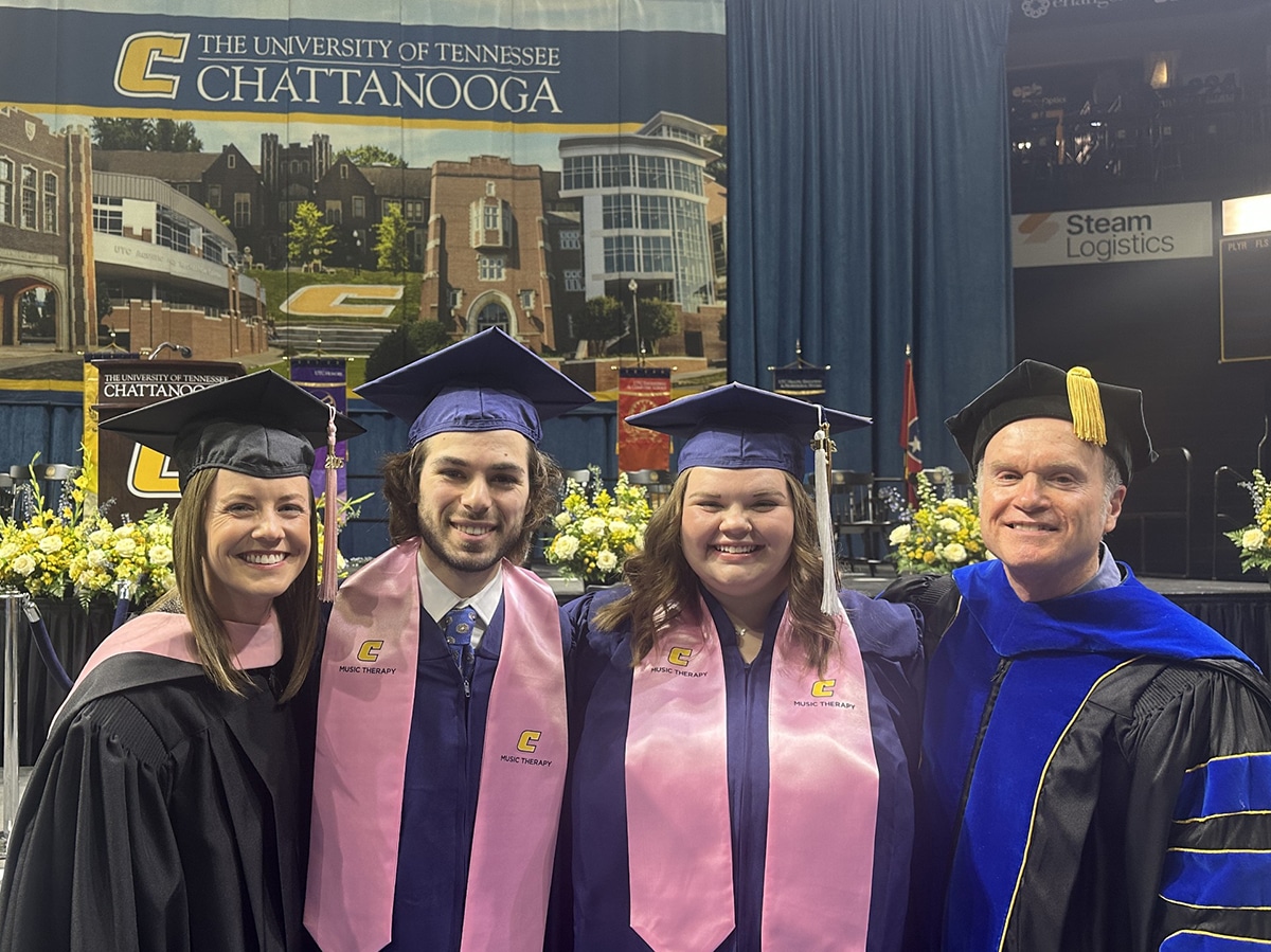 Four people in academic regalia standing shoulder to shoulder at commencement